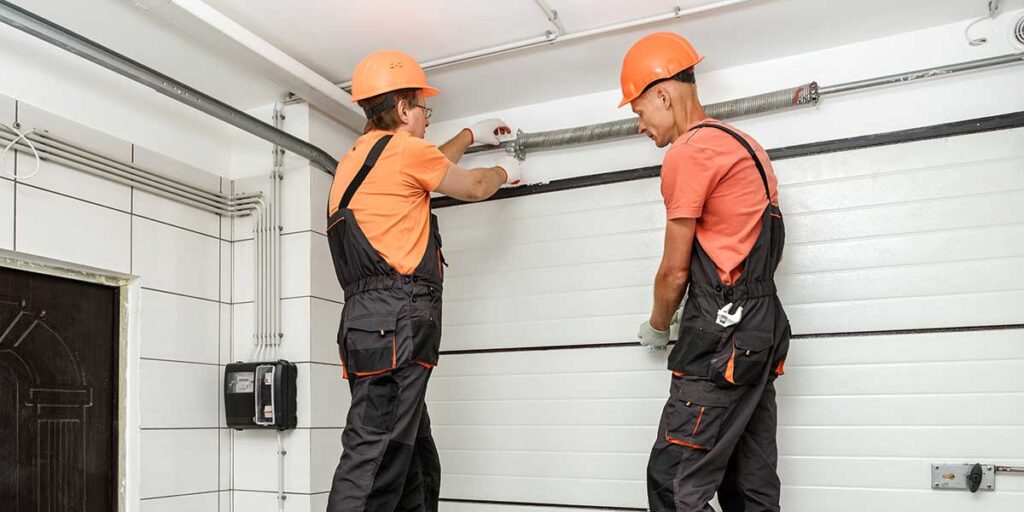 Two technicians wearing hard hats inspect and tune a residential garage door system following a maintenance checklist.